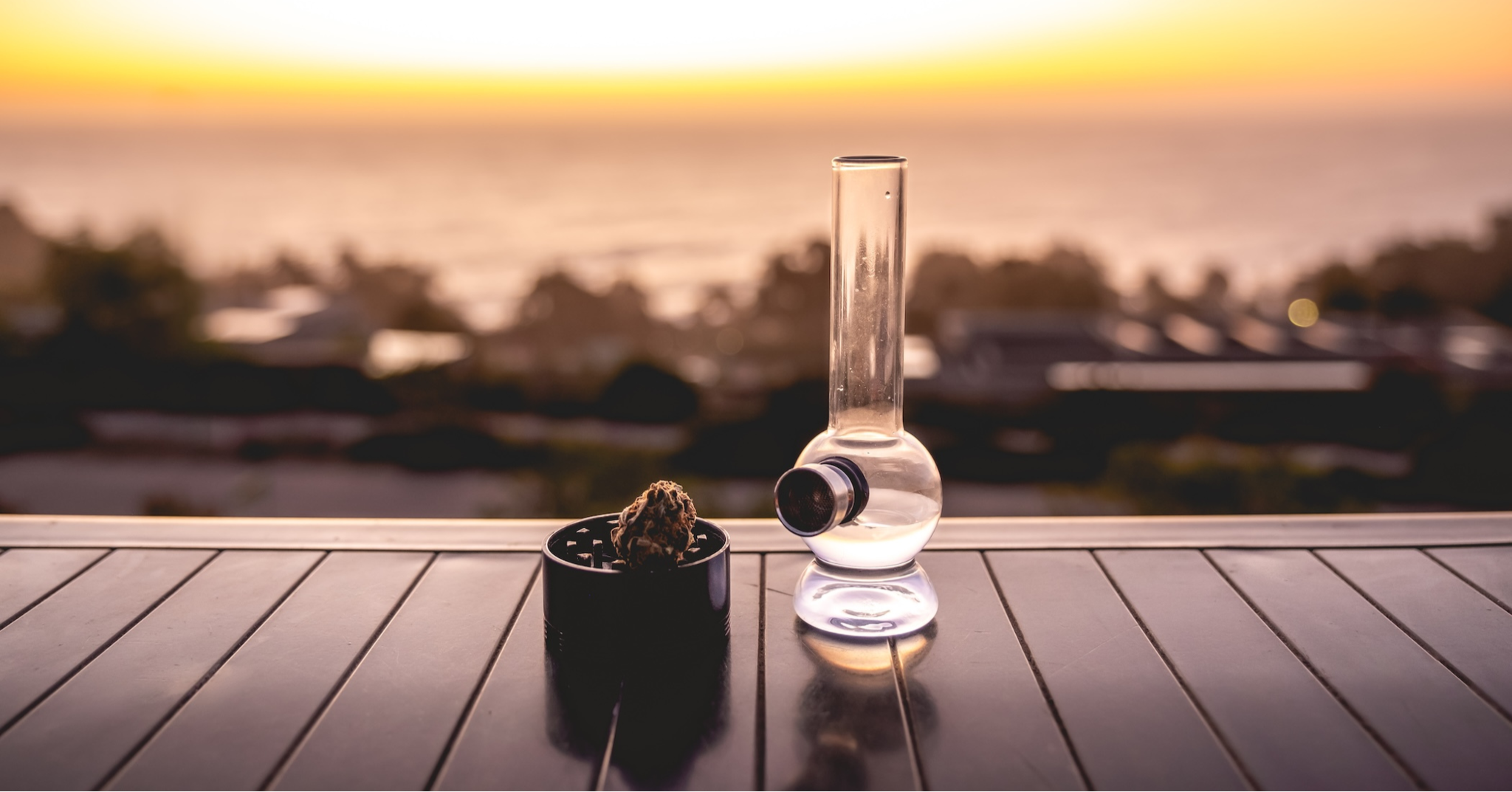 Bong and grinder filled with cannabis on a table in sunset]
