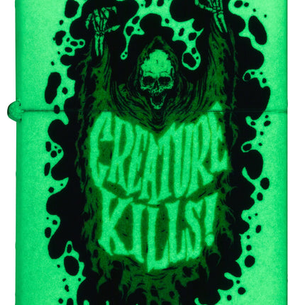 The Zippo 'Creature Kills!' lighter glowing bright neon green in the dark, highlighting the skeletal figure and text through its glow-in-the-dark finish.